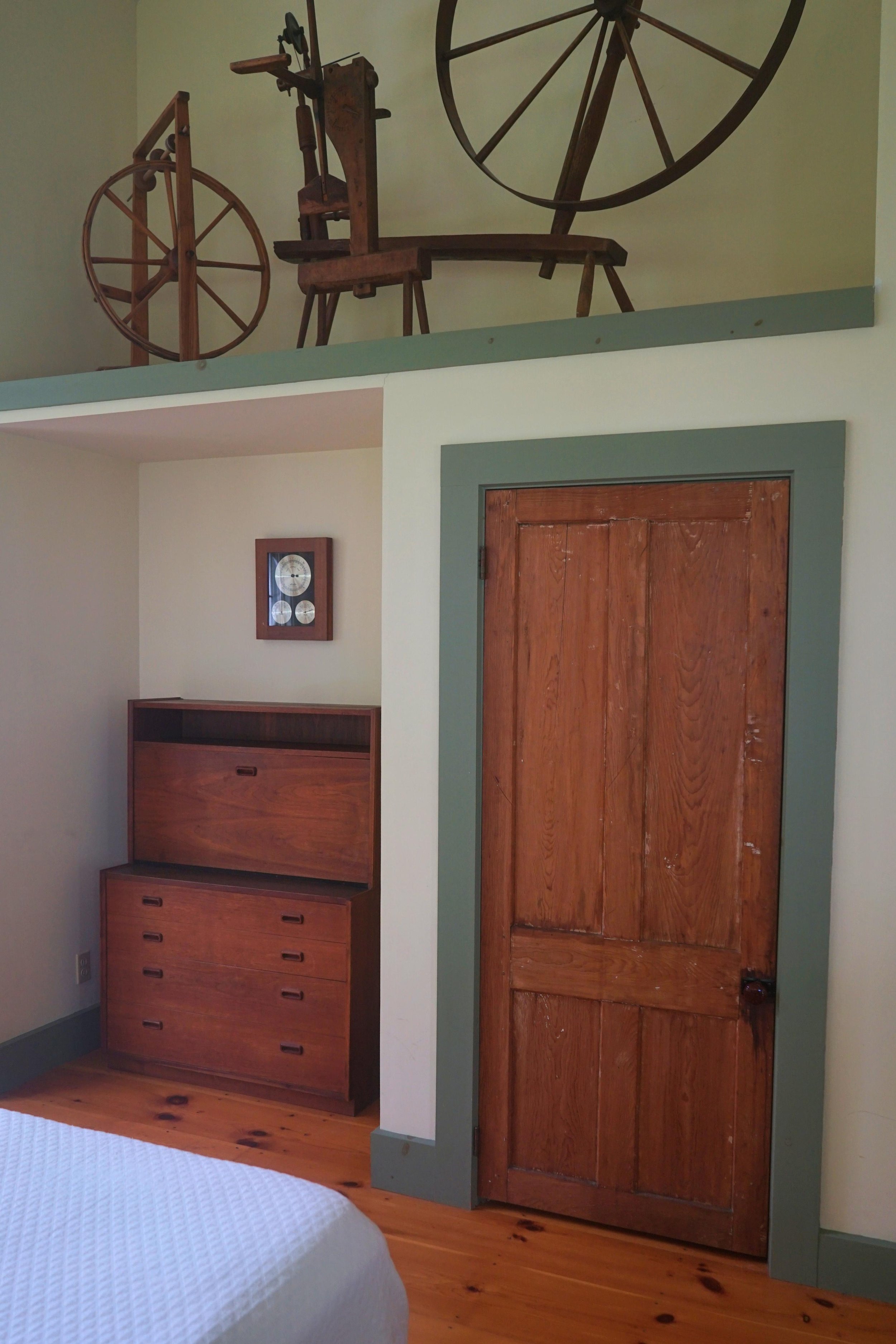 Room with wooden door, wooden dresser, and wall clock, with a display of antique spinning wheels on the upper level.
