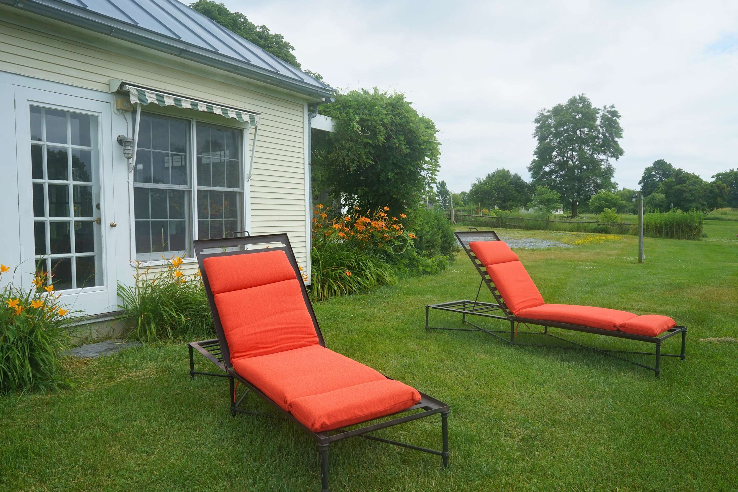 Two orange outdoor lounge chairs with cushions on a green lawn next to a white house with yellow siding, flower bushes, and a tree in the background.