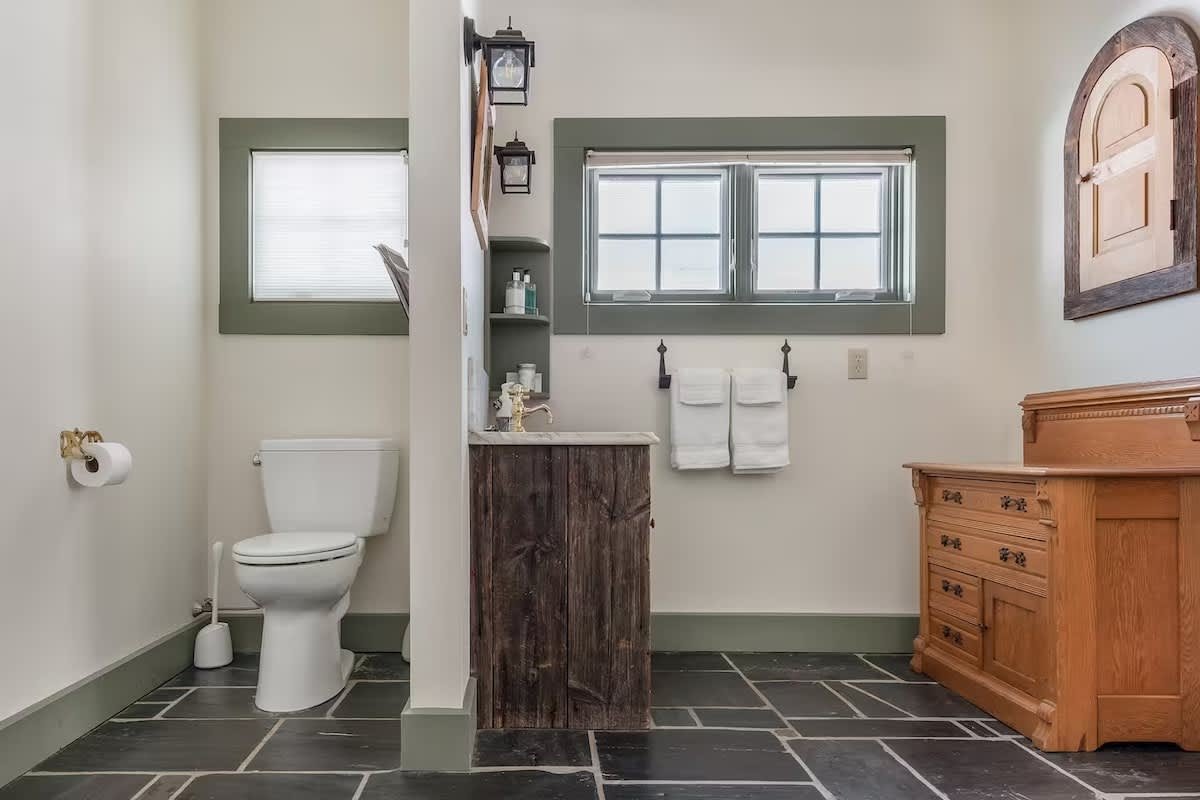 Bathroom with a toilet, a sink with a rustic wood cabinet, a wooden dresser, and two side-by-side windows with green trim.