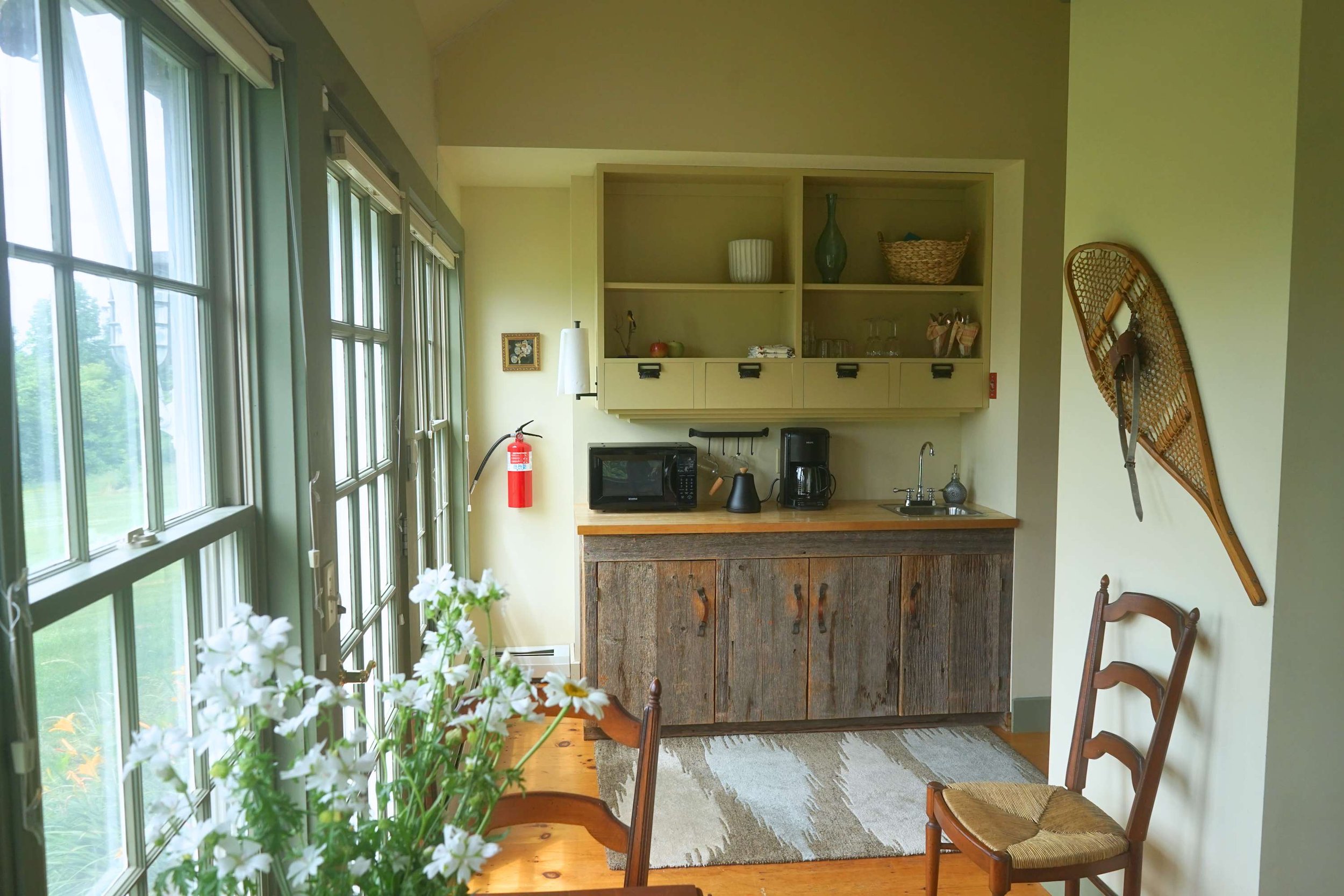 Cozy kitchen corner with wooden cabinets, black microwave, coffee maker, and toaster, next to a small sink. Large windows with greenery outside, a pair of wooden chairs, a snowshoe hanging on the wall, and a patterned rug on a wooden floor.