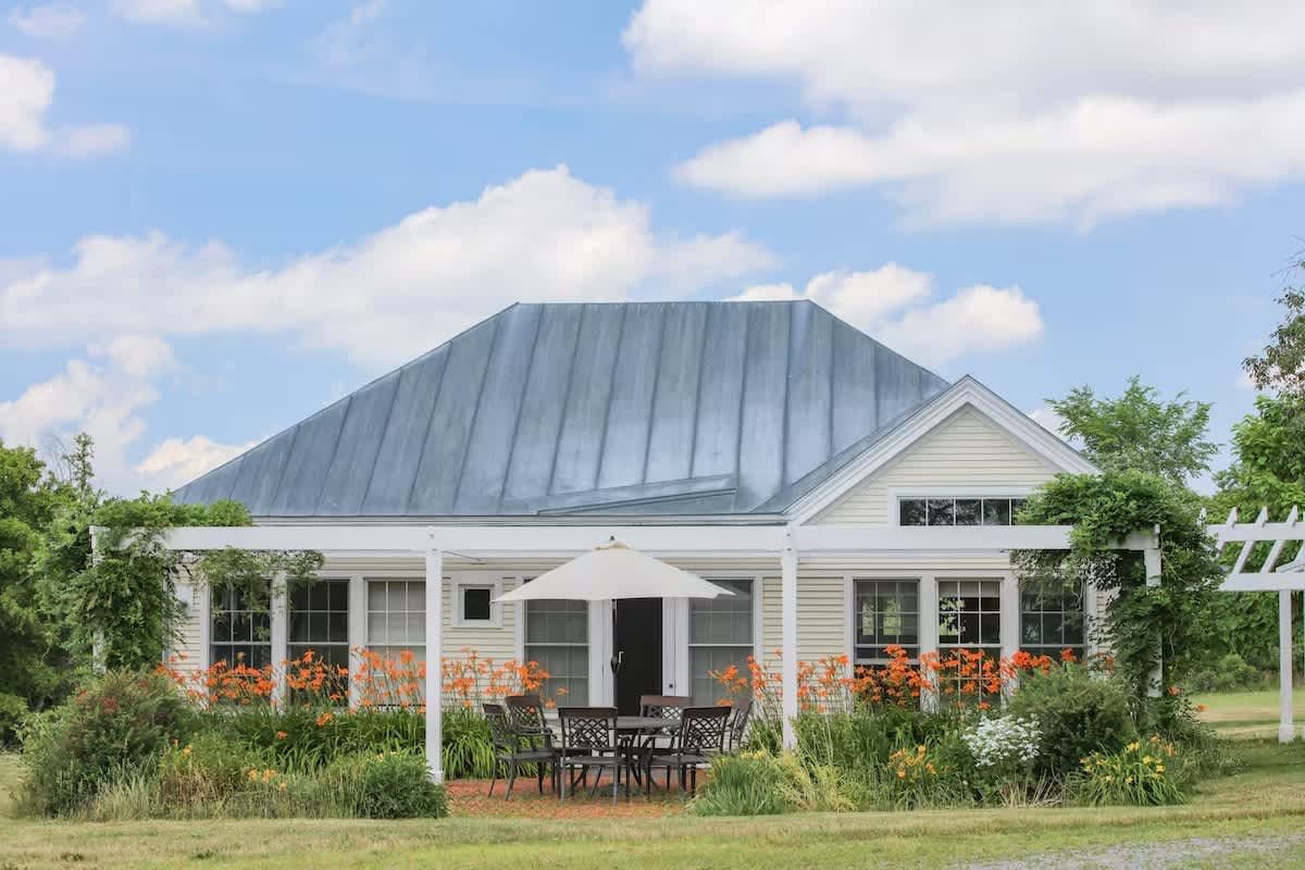 Guest house surrounded by a garden with orange and yellow flowers, a patio with a table and chairs, and a white umbrella, under a blue sky with clouds in idillic Addison County Vermont.
