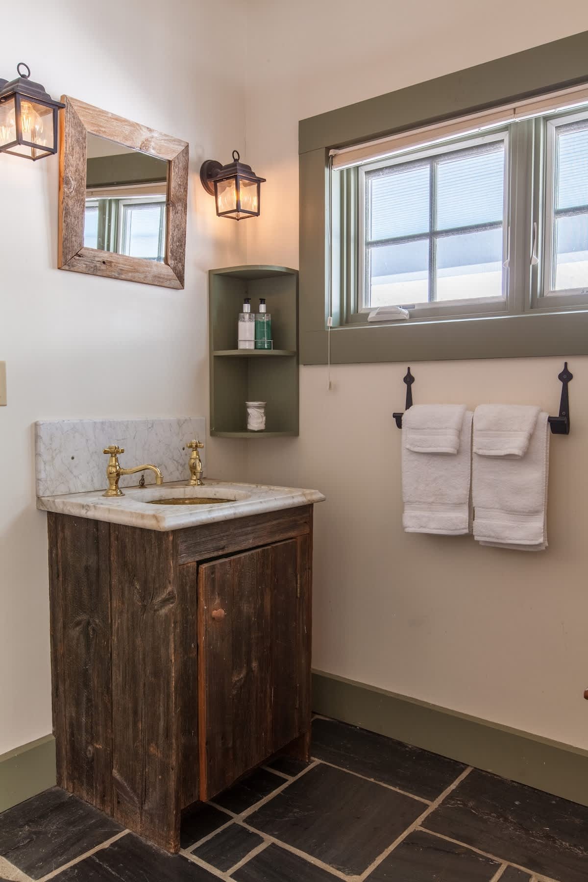 A rustic bathroom with a wooden vanity, a marble sink, gold faucet, green shelves, and towels hanging on a black metal rack. A window with blinds and two wall lanterns are visible.