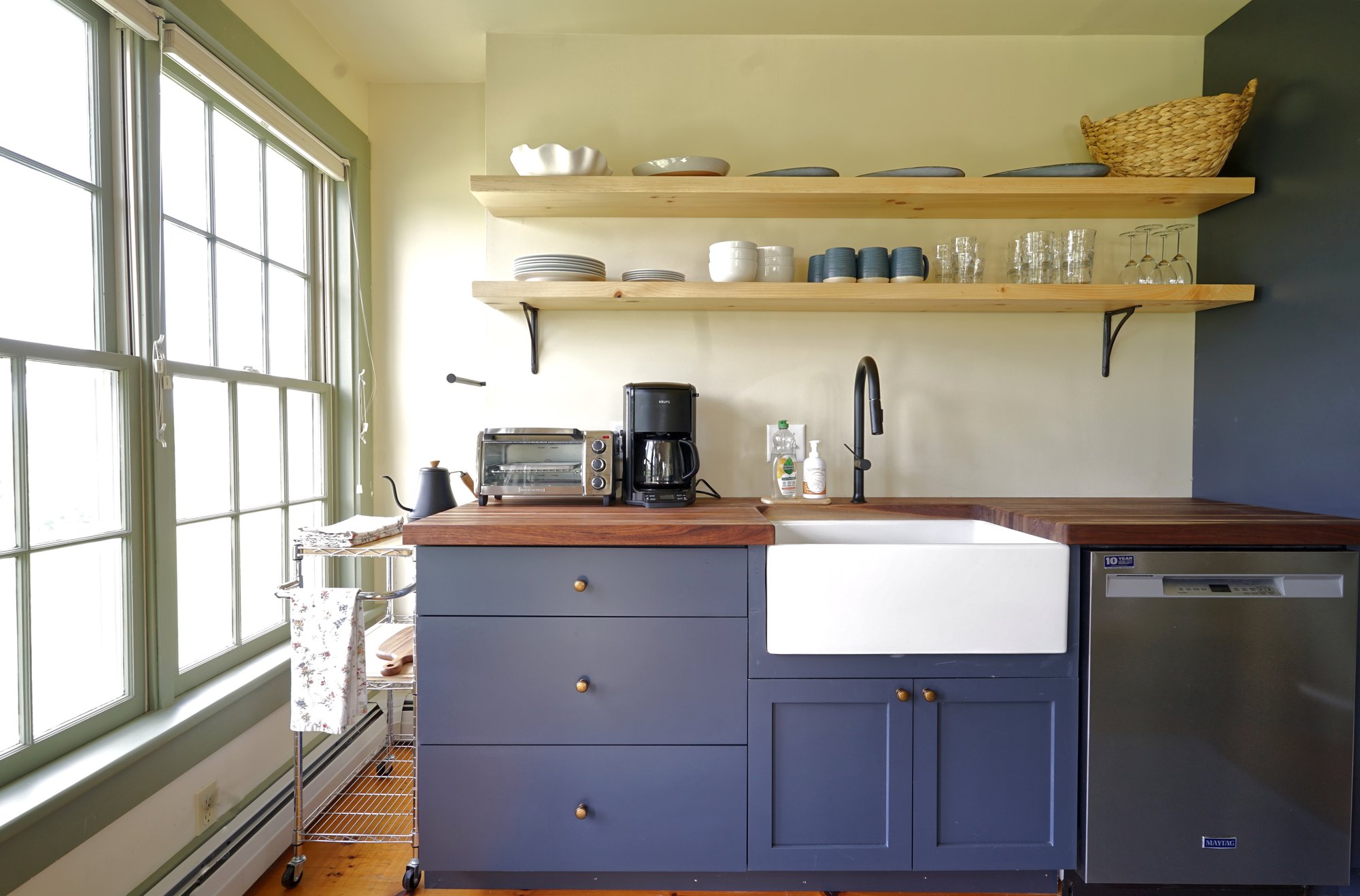 Kitchen counter with coffee maker, toaster oven, farmhouse sink, and dishwasher beneath open shelving and windows.
