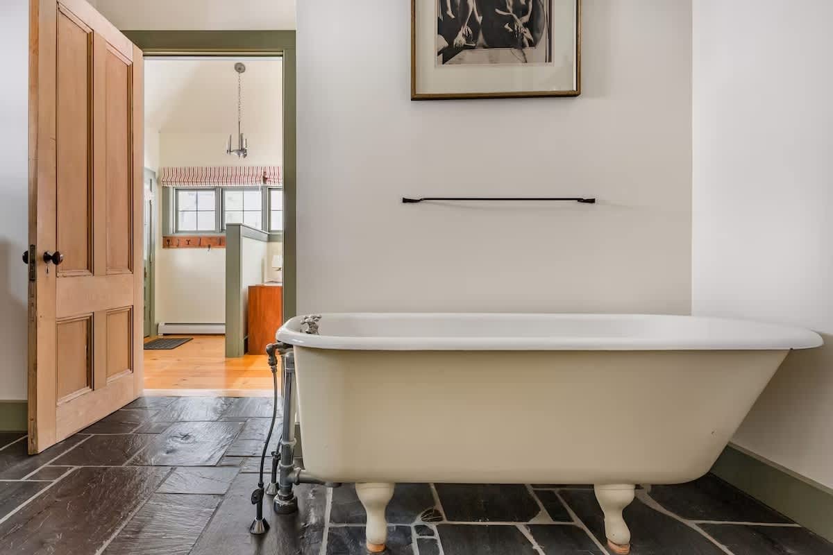 A white clawfoot bathtub in a bathroom with black stone tile flooring, an open wooden door leading to a room with a window, a framed photograph on the wall, and a black towel bar.