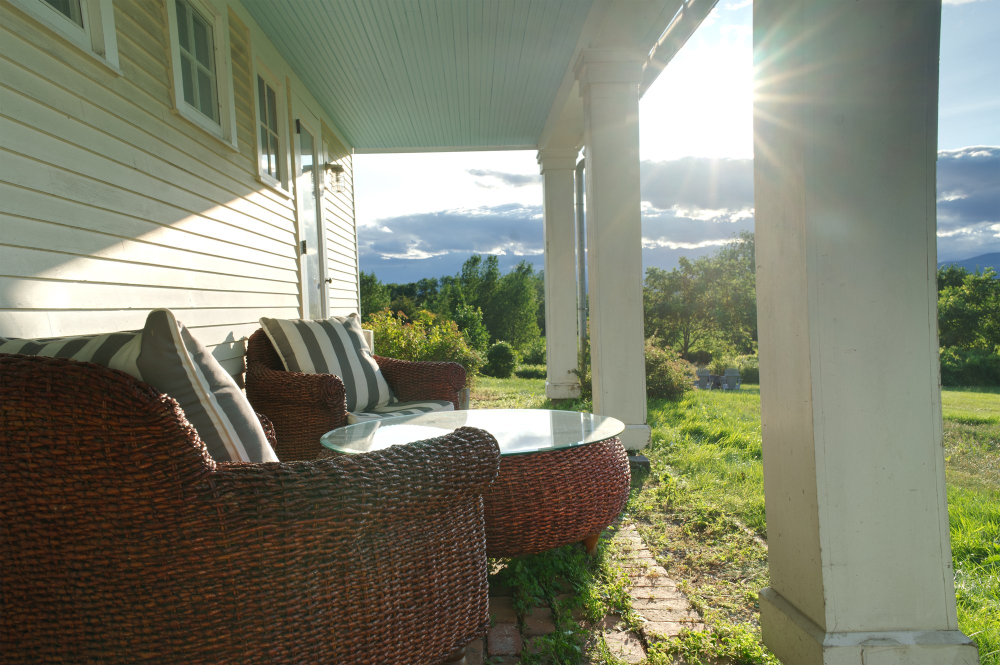 Wicker chairs with striped pillows and a glass-top table on the covered porch, with the sun setting over the mountains beyond.