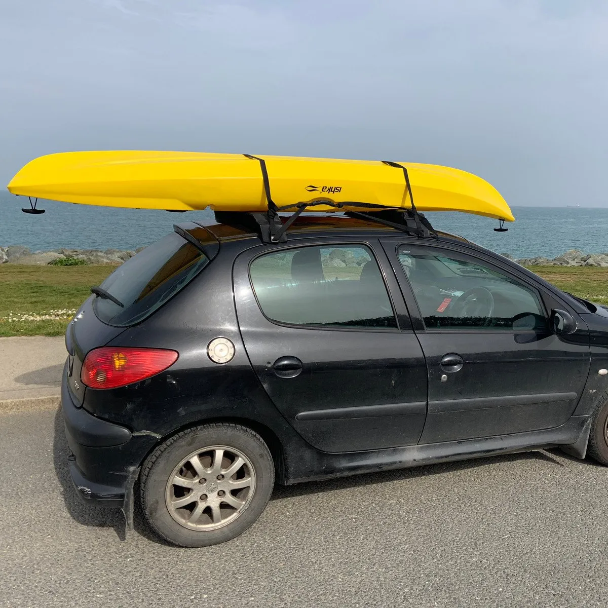 A kayak strapped onto soft roof pads on top of a small hatchback by the water